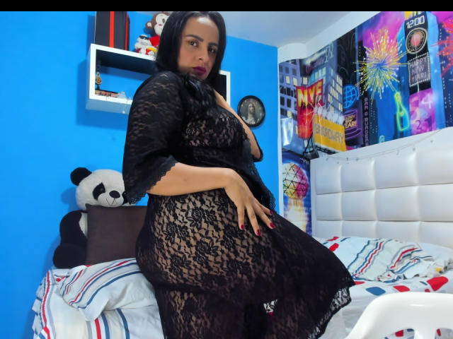 PamelaRouss in un abito di pizzo nero, in piedi sul letto, che mostra il sedere, il seno di media taglia e le unghie rosse, con un peluche a forma di panda nella stanza con le pareti blu..