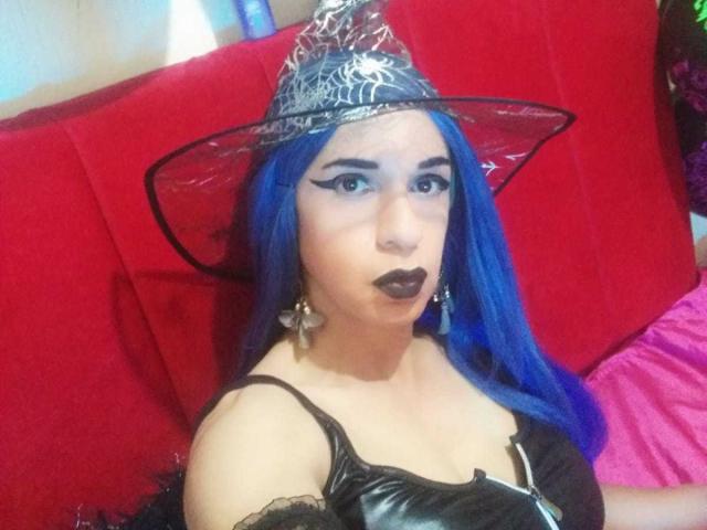 Bellanida with blue hair, black lipstick, and a spiked witch hat, wearing a black leather top, sitting on a red couch. Her tits are visible, and she's looking at the camera.