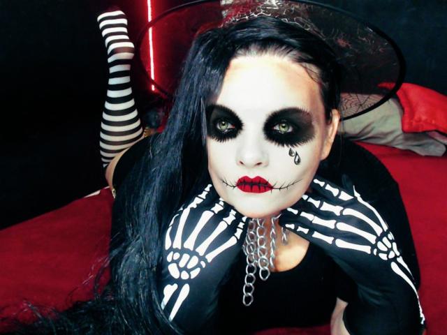 Andrea Sharlin, avec du maquillage blanc sur le visage, du maquillage noir autour des yeux, des lèvres rouges, des gants noirs en forme de squelette, de longs cheveux noirs, portant un haut noir et un collier en chaîne, sur un lit rouge..