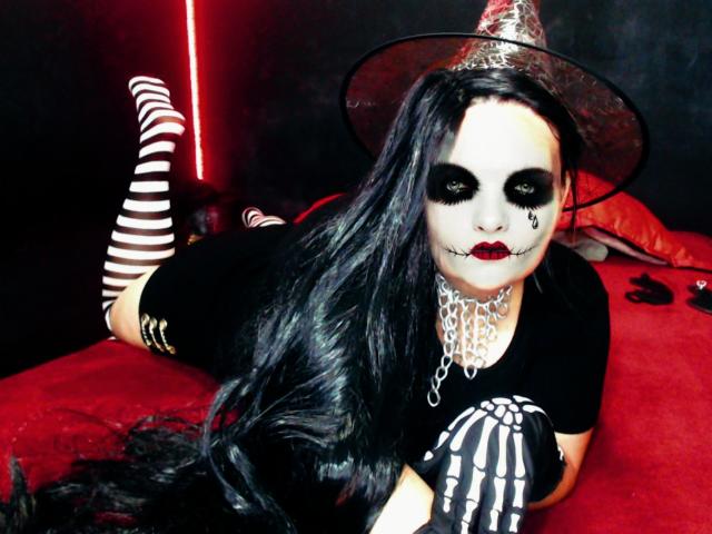 AndreaSharlin, avec du maquillage pâle sur le visage, du rouge à lèvres noir, de longs cheveux noirs, un chapeau de sorcière, des bas rayés hauts, une chemise noire, un gant squelette, allongée sur un lit rouge, portant un collier en chaîne blanche..