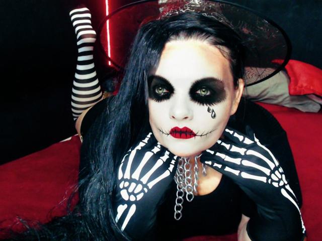 AndreaSharlin met witte gezichtsbeschildering, zwarte oogmake-up, rode lippen, zwarte handschoenen in skeletvorm, een zwarte jurk, een ketting, gestreepte kniekousen, een zwarte hoed, lang zwart haar, liggend op een rode bed..