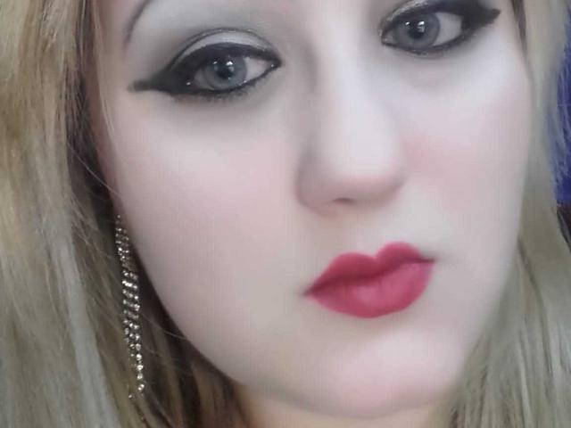 SeductiveBodyX has pale skin, blue eyes with thick black eyeliner, and red lipstick. Her blonde hair is down, and she's wearing silver earrings. Her face is slightly blurred.