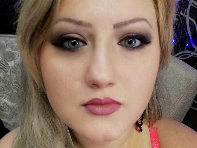 SeductiveBodyX has blue eyes, black smokey eye makeup, pink lipstick, and blonde hair. She's wearing a neon pink strap, and a black beaded necklace is visible.