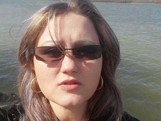SeductiveBodyX has brown eyes, wearing dark sunglasses, with light brown hair, small gold hoop earrings, and pink lips, standing by a greenish water body.