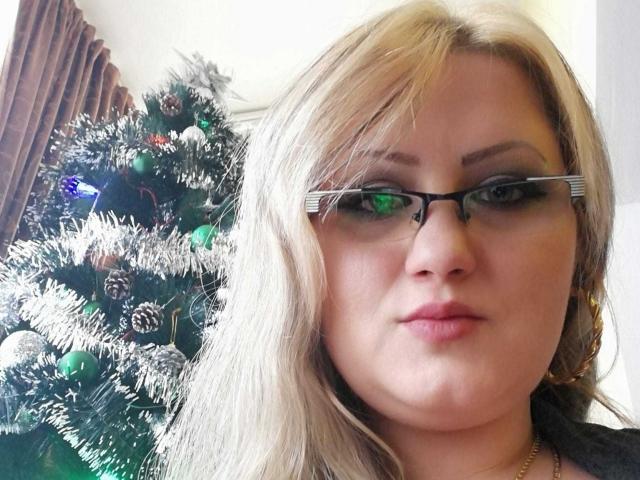 SeductiveBodyX with blonde hair, glasses, and pink lipstick, standing next to a decorated Christmas tree, showing a serious face. Boobs not visible, pussy not visible.