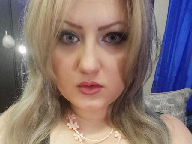 SeductiveBodyX with blonde hair, blue eyes, pink lipstick, and a flower necklace, staring at the camera with a slightly parted mouth, showing a cute and sexy face.