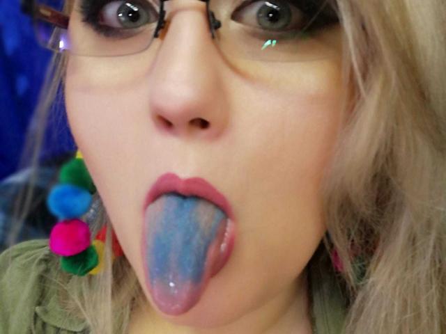 SeductiveBodyX's face with blue-tongued open mouth, glasses, and blonde hair, wearing colorful earrings, has wide eyes looking forward. Her nose is centered in the shot.