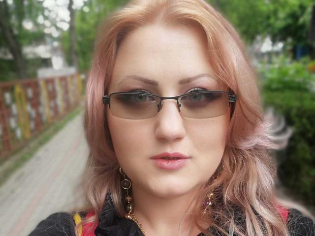 SeductiveBodyX with pink hair, glasses, and pink lipstick, wearing a black shirt with gold earrings, looking straight at the camera, showing a confident, slightly smug expression.