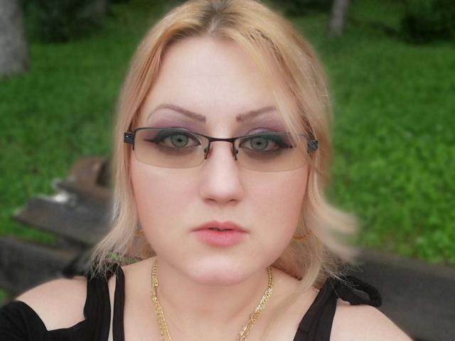 SeductiveBodyX has blonde hair, green eyes, and wears black glasses with green eye makeup. She's looking directly at the camera with a neutral expression. Her tits are partially visible.