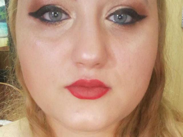 SeductiveBodyX has blue eyes with black eyeliner, red lipstick, and pale skin. Blonde hair, slightly parted, with a neutral expression. Close-up shot, face-focused.