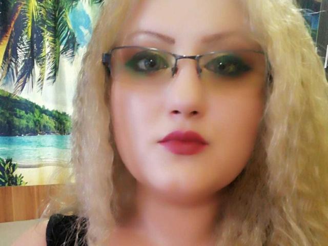 SeductiveBodyX has curly blonde hair, green eyeshadow, and red lipstick, wearing glasses, with a serious expression, and a black top, in a blurry photo.