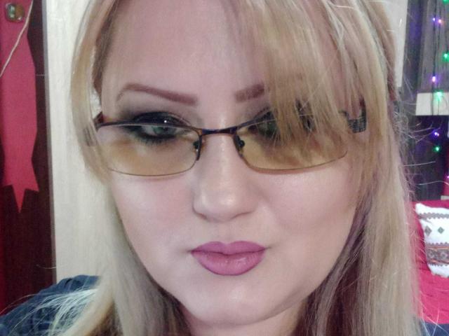 SeductiveBodyX has blonde hair, glasses, and pink lipstick, with a slightly pouty face. Her green eyes are partially hidden by her bangs. She's wearing a dark shirt.