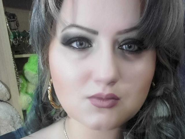 SeductiveBodyX has blue eyes, heavy black eyeliner, and full pink lips. She has wavy gray hair, gold earrings, and a slight smile. Her face is centered, looking at the camera.
