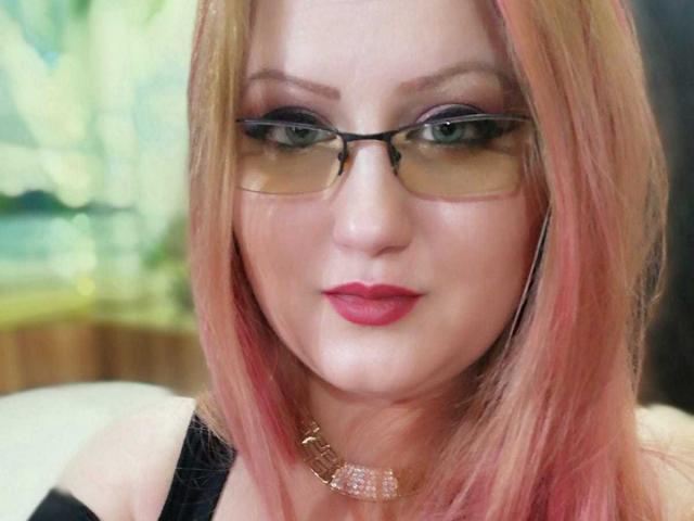 SeductiveBodyX has pink and blonde hair, wears glasses, and has pink lipstick with a jeweled choker. Her green eyes are slightly squinted, giving a cute, confident look.