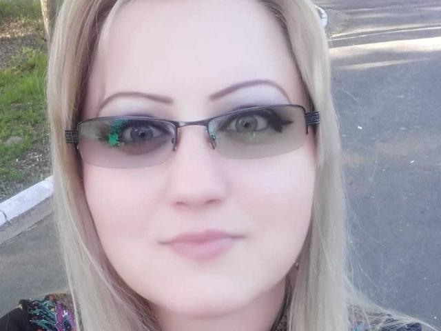 SeductiveBodyX has blonde hair, green eyes, and wears black glasses with thick frames. Her pink lips are slightly curved in a subtle smile. She's standing outside on a sunny day.