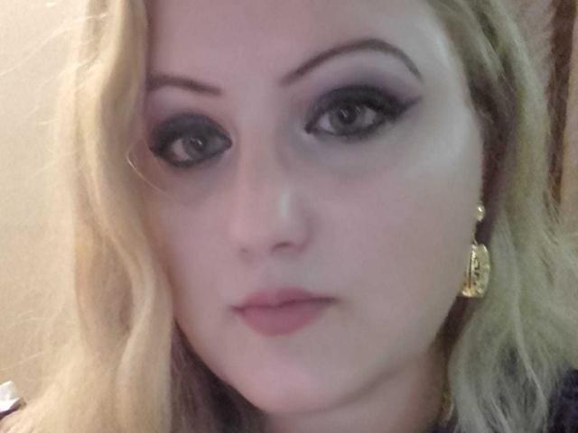 SeductiveBodyX has pale skin, blonde hair, and is wearing dark eyeliner with bold eyebrows. She has gold earrings and pink lipstick. Her eyes are looking directly at the camera.
