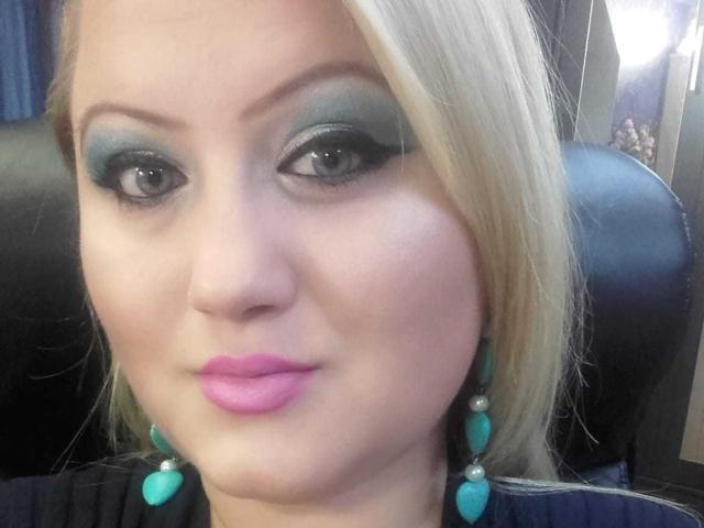 SeductiveBodyX has blonde hair, green eyeshadow, black eyeliner, pink lipstick, teal earrings, and a light skin tone, looking at the camera with a slight smile.