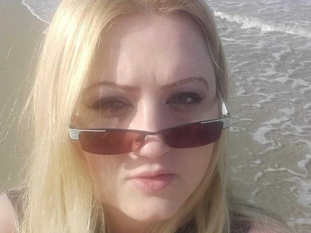 SeductiveBodyX has blonde hair, wearing brown sunglasses, with fair skin, and a slight pout. Her tits are partially visible under a shirt, standing by the ocean.