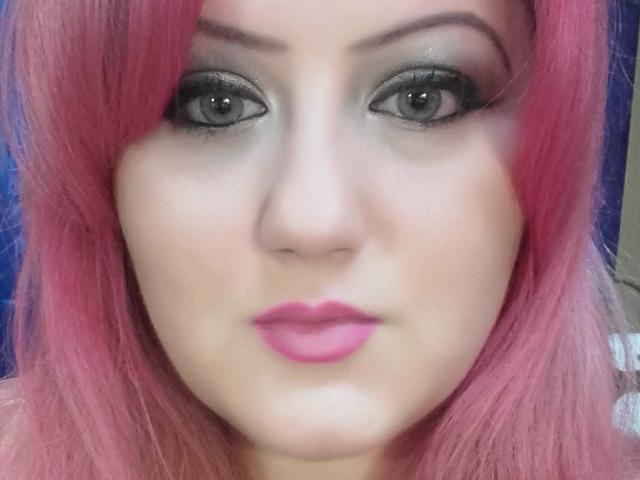 SeductiveBodyX has pink hair, green eyes with heavy black eyeliner, and pink lipstick. She's staring directly at the camera with a neutral expression. Her skin is fair.