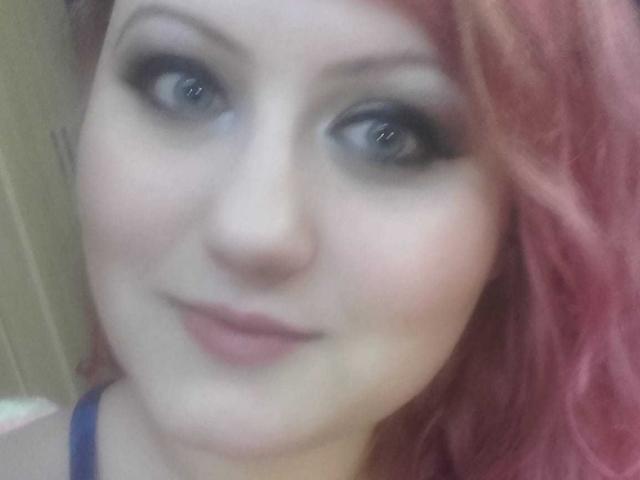 SeductiveBodyX has pale skin, red wavy hair, and dark eyeliner with smoky eyes. She's smiling slightly with pinkish lips, showing a subtle, sexy look.