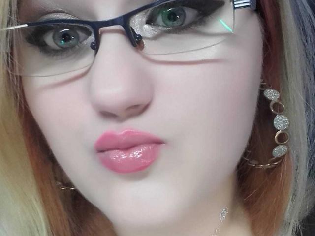 SeductiveBodyX has pale skin, green eyes, and glossy pink lips. She wears black-rimmed glasses and gold earrings with circular studs. Her blonde hair has orange highlights.
