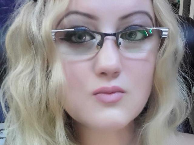 SeductiveBodyX has blonde wavy hair, green eyes, and wears black rectangular glasses with reflective green and white light. Her pink lips are slightly parted.
