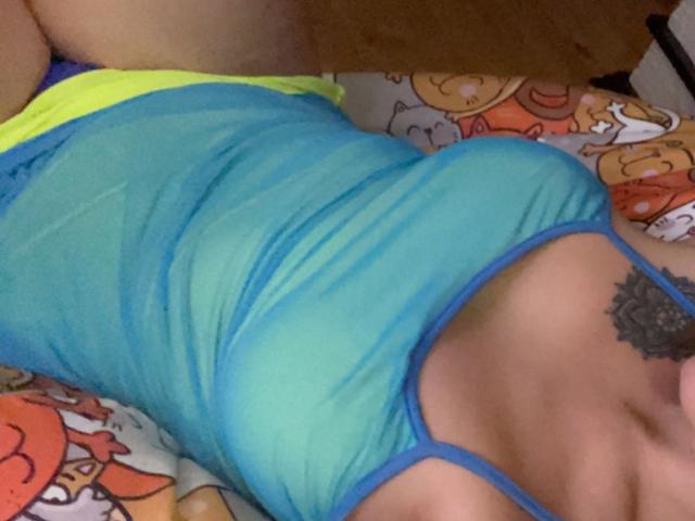 Misstyk in a blue, sheer, spaghetti-strap top, lying on a colorful bed, with yellow shorts, showcasing her tits and a black tattoo on her chest.