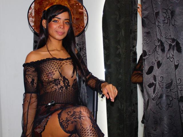 MelisaLover, con cabello oscuro, vestida con un vestido negro de encaje transparente que deja ver sus senos y vulva, sentada con un halo, cortinas verdes y patrones florales en el fondo..
