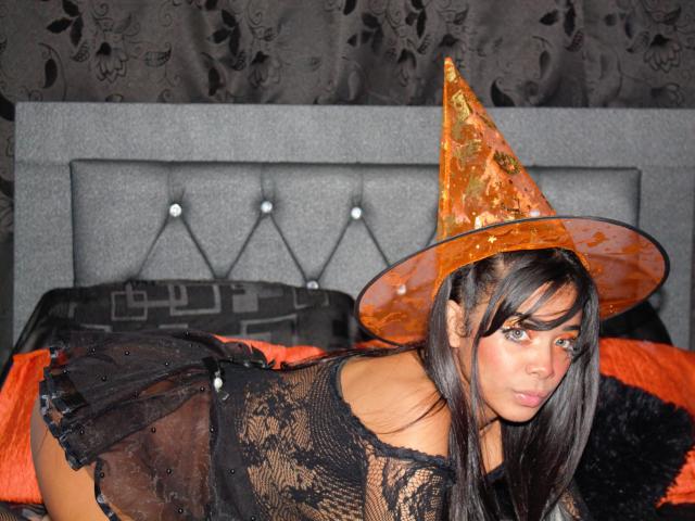 MelisaLover with long black hair, wearing a transparent lace black dress, orange witch hat, and looking over her shoulder, showing her ass. She's on a grey bed with orange pillows.