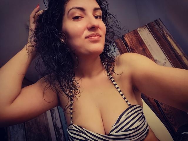 AddictedToUTS with curly black hair, smiling, wearing a striped black and white bikini top, showing her big tits, holding her hair, in a dimly lit room.