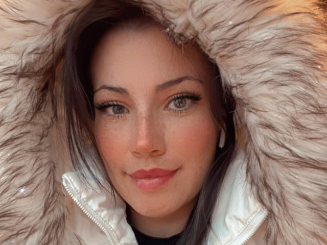 Liliysweet mit flauschigem Winterfell, grauen Augen und rosafarbenen Lippen, die leicht lächelt. Sie trägt kleine, weiße Ohrringe und hat ein süßes, mit Sommersprossen übersätes Gesicht. Braune Haare, dezentes Make-up..