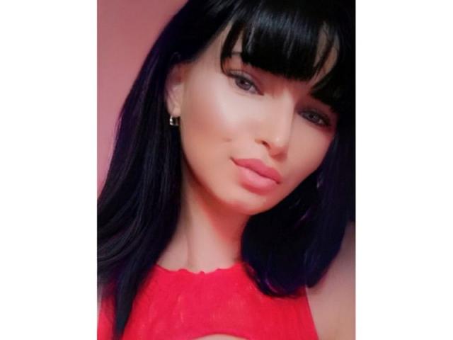 Allexandra Terri tiene el cabello negro oscuro con flequillo, lleva una blusa roja de encaje y tiene los labios rosados con un ligero bostezo. Está mirando a la cámara con una expresión adorable..
