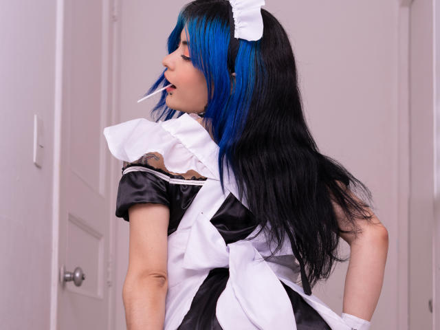 CandiceValo, avec ses cheveux bleus et noirs, portant une tenue de femme de ménage noire et blanche, a ses fesses tournées vers la caméra, et regarde par-dessus son épaule, avec un cure-dent blanc dans la bouche..