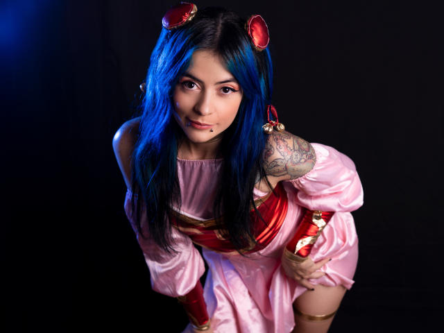Candice Valo, avec ses cheveux bleus, sa robe rose, ses brassards rouges et son serre-tête orné de cornes rouges, penchée en avant, montrant un tatouage sur son bras droit et un petit piercing sur la lèvre..