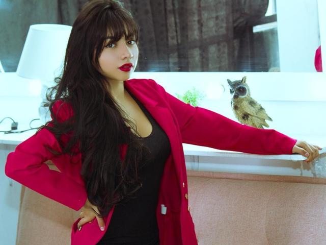 SakuraMiomi with long brown hair, red coat, black top, and red lipstick, leaning on a counter with a small owl on it, hand on hip, looking at camera.