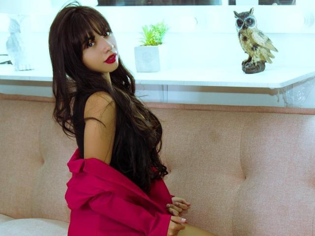 SakuraMiomi mit langen braunen Haaren, rotem Lippenstift und einem pinken Kleid, sitzt auf einem pinken Sofa, die pinke Jacke ist ausgezogen, wodurch ihre nackten Schultern und ihr Rücken sichtbar sind, neben einer Eulenstatue..