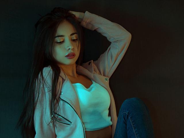 SakuraMiomi, com cabelos longos e pretos, vestindo uma jaqueta rosa, um cropped branco e jeans azuis, tem olhos dourados e uma mão no cabelo, sentada com as pernas levantadas..
