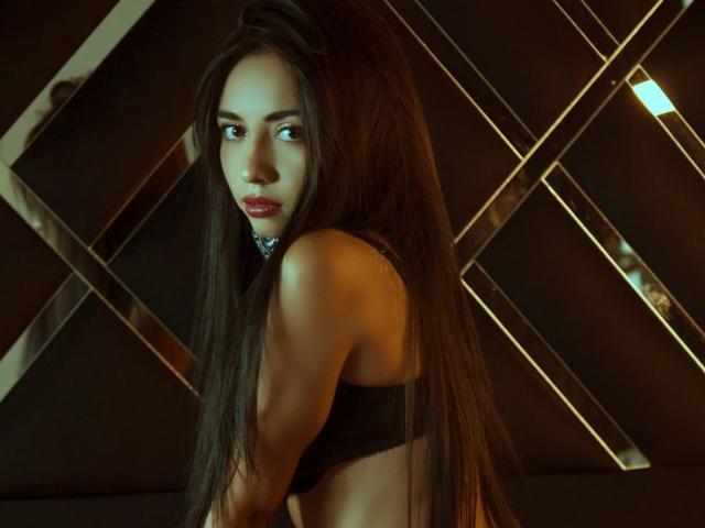 SakuraMiomi, avec de longs cheveux bruns, des yeux foncés, du rouge à lèvres rouge, un soutien-gorge noir, regardant par-dessus son épaule, montrant ses fesses fermes et son dos mince, dans une pièce faiblement éclairée, avec des accents dorés..