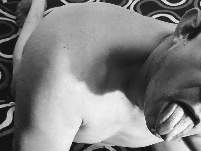 Fotografia in bianco e nero della parte superiore del corpo di Horgasmik, muscolosa e senza camicia, che mostra una spalla forte e definita e una bocca leggermente aperta con la lingua visibile. La testa di Horgasmik è inclinata verso destra..