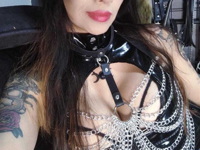 ScarlettRoseX indossa un'imbracatura in lattice nero, con dettagli in catena metallica, e ha i seni scoperti. Ha tatuaggi sul braccio destro ed indossa rossetto rosso. Il suo viso è parzialmente visibile..