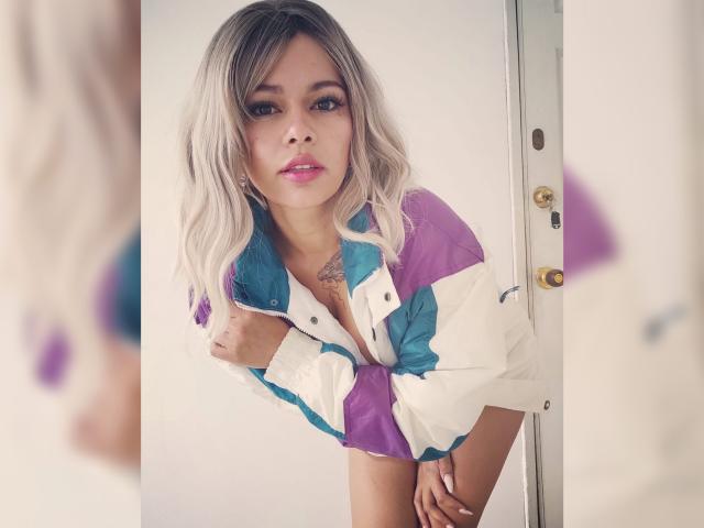 Daria Dawson, com cabelo loiro, vestindo uma jaqueta colorida e abotoada, inclinada ligeiramente para frente, com as mãos na porta, mostrando uma pequena tatuagem no peito, lábios rosados, olhando para a câmera..