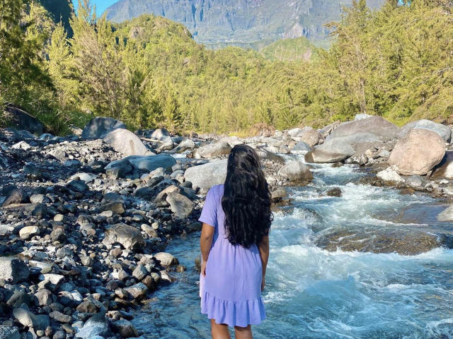 StormyKiss è in piedi con un abito viola e lunghi capelli neri, di fronte a un fiume roccioso con acqua limpida e alberi verdi, con la schiena rivolta verso la telecamera..