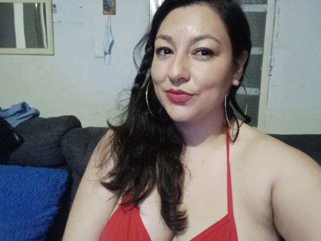 SakotoDika mit mittelgroßer Brust, trägt ein rotes Halstopp, hat schwarzes, welliges Haar, trägt Ohrringe im Reifenstil und roten Lippenstift, sitzt auf einem grauen Sofa und lächelt sanft..