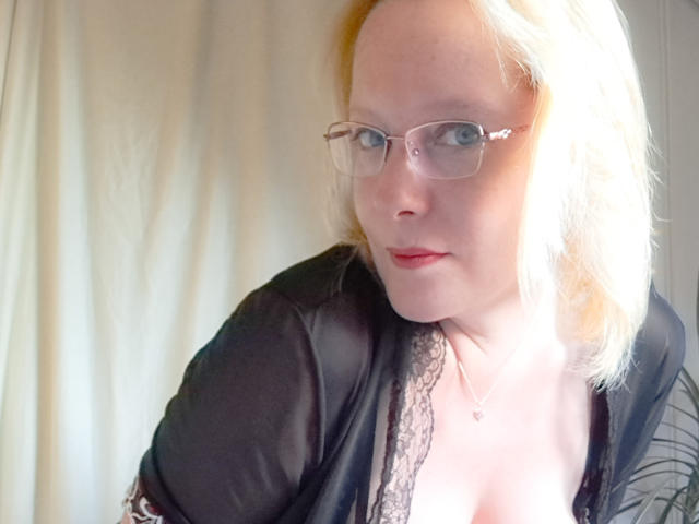 EleanaRose with blonde hair, glasses, and red lipstick, wearing a black lace-trimmed top, showing her small tits. She's looking at the camera with a slight smile.
