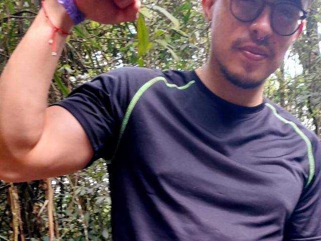 Darko, mit Brille und Nasenring, spannt seinen Arm in einem schwarzen Hemd mit grünen Akzenten, hält ein Blatt in der Hand, trägt ein violettes Armband und eine rote Perlenkette..