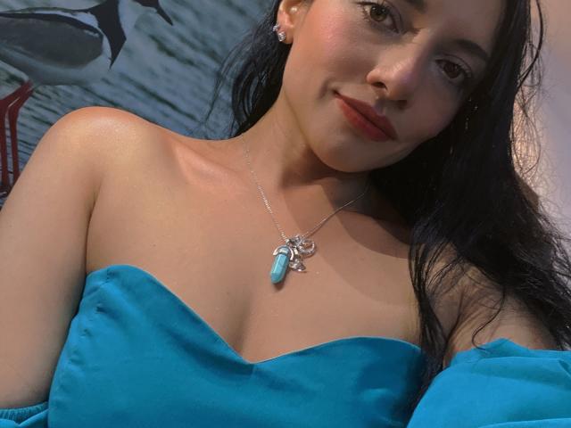 AryredStars com seios médios, vestindo uma blusa azul sem alças, um colar de prata com um pingente de turquesa e brincos, sorrindo suavemente, cabelos pretos longos, pele clara, maquiagem discreta..