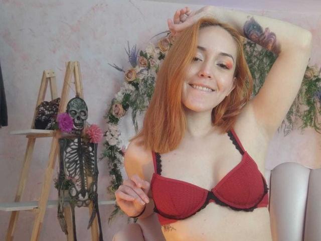 MollyMarmalade con un sujetador rojo, mostrando sus pechos, sonriendo, con el brazo levantado, tatuajes visibles, decoración de calaveras en una escalera, una guirnalda floral, una pared rosada y una silla en el fondo..