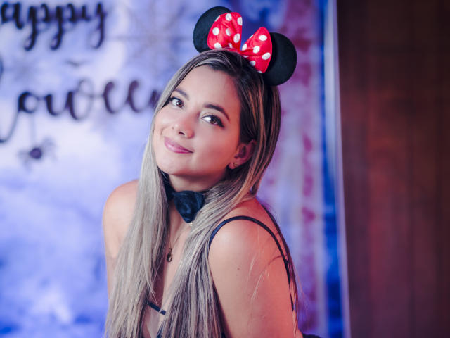 AbrilClark with long blonde hair, wearing black strap dress, Minnie Mouse ears with red polka-dot bow, black bow tie, smiling at camera, blue and purple background, cute.