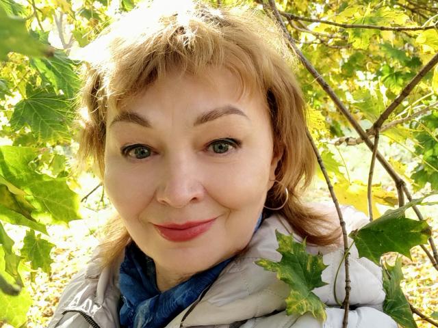 A MilaMils tem um rosto adorável, com olhos verdes, cabelo ondulado castanho claro e batom rosa. Ela está vestindo uma jaqueta branca e sorrindo em um ambiente externo ensolarado e com muitas folhas..
