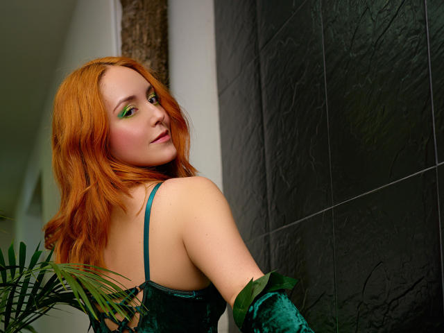 MollyMarmalade with red hair, green eyes, green velvet dress, looking over shoulder, green leaf in hand, black textured wall, palm leaves in foreground, soft smile, cute face.